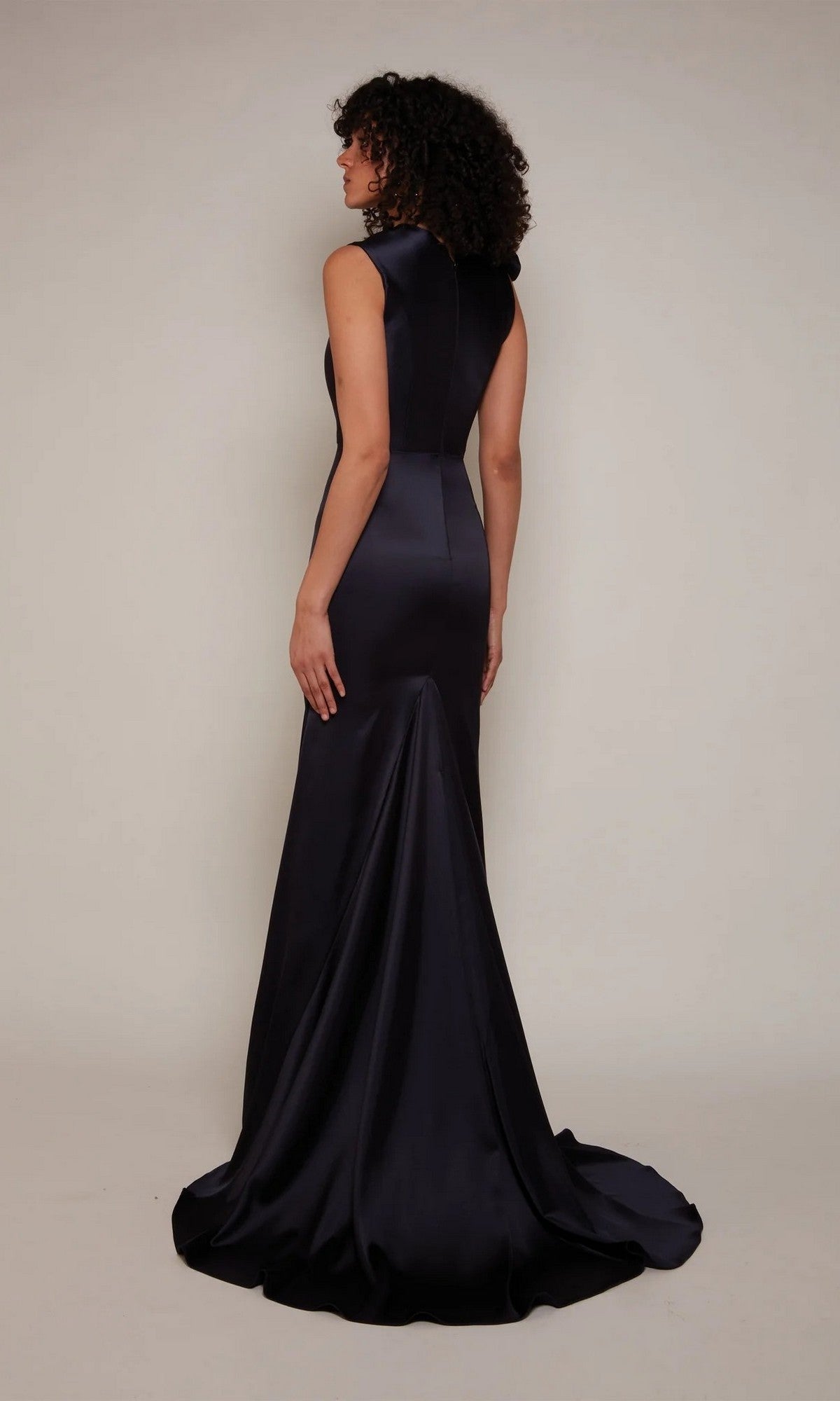A woman stands facing away, wearing the Long White Dress: Alyce 7067—a sleeveless, floor-length satin sheath gown with a subtle sweep train, set against a plain background.